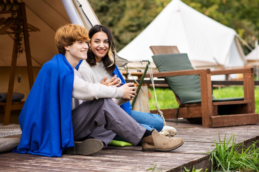 A couple sitting under a blanket on a tent terrace, holding cups and smiling at glamping. Lush forest around