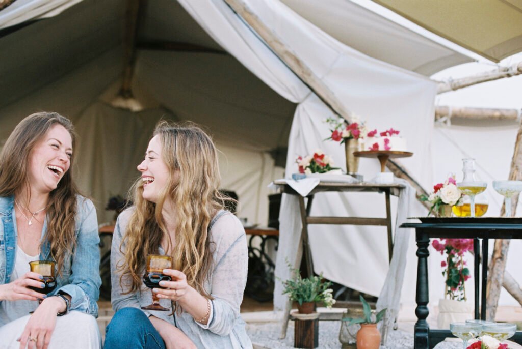 Two smiling women sitting outside a tent in a desert, enjoying a glass of wine.